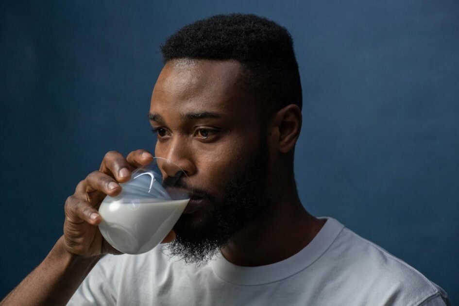 A serious man in a white t-shirt drinks milk from a glass against a dark blue background.